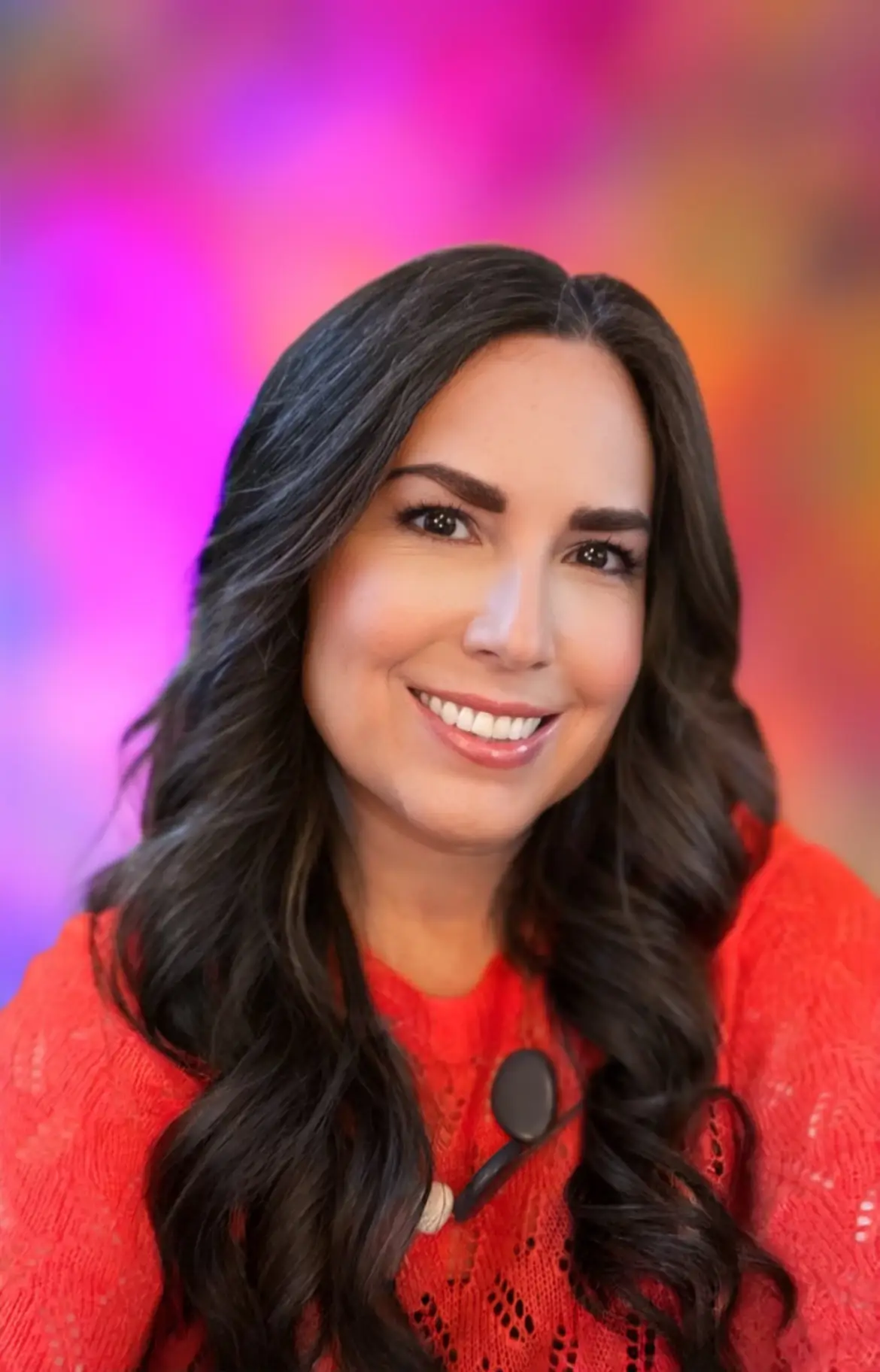 Smiling woman with long, wavy dark hair, wearing a red textured top, in front of a colorful blurred background with pink, purple, and orange tones.