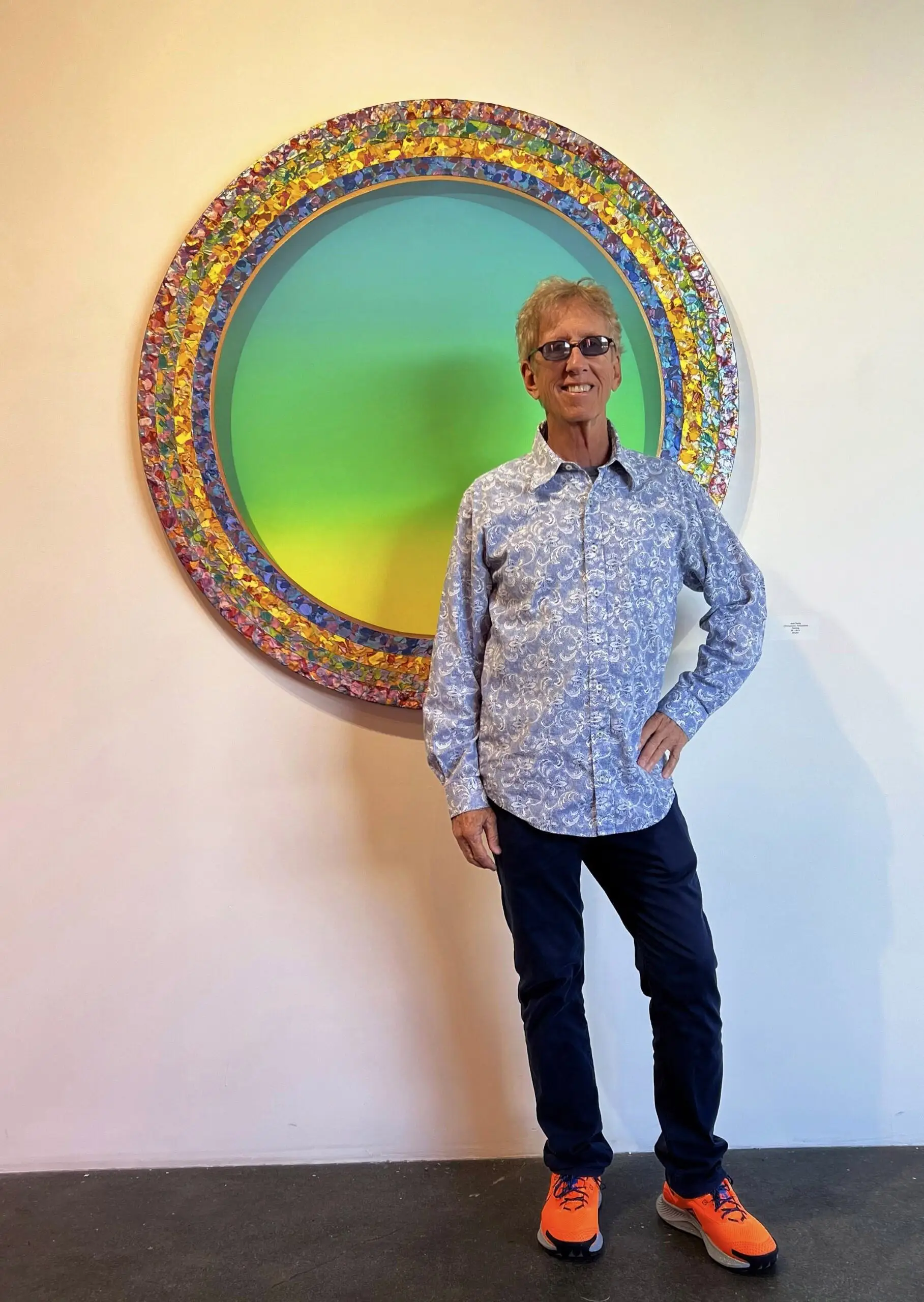 Man with short gray hair and glasses, standing in front of a large circular artwork with a green and blue center surrounded by colorful textured rings, wearing a light blue patterned shirt and orange sneakers.