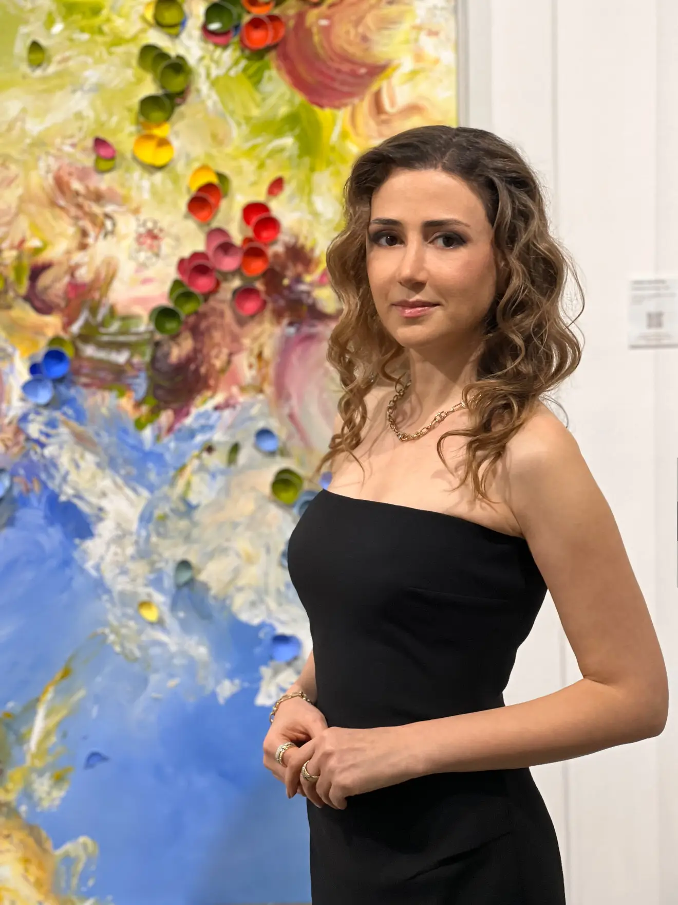Woman with long curly hair wearing a strapless black dress and gold jewelry, standing in front of a colorful abstract painting featuring swirling textures and rolled paper elements.