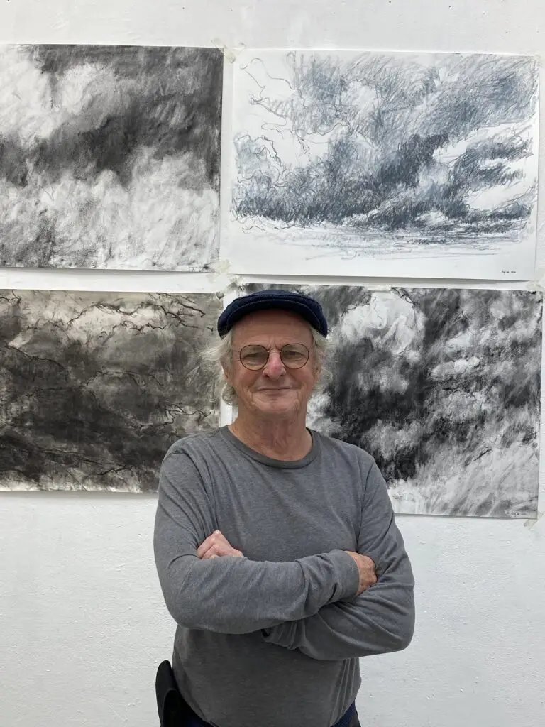 Older man with glasses and a blue cap, wearing a gray long-sleeve shirt, standing with arms crossed in front of four black and white abstract landscape drawings taped to a white wall.