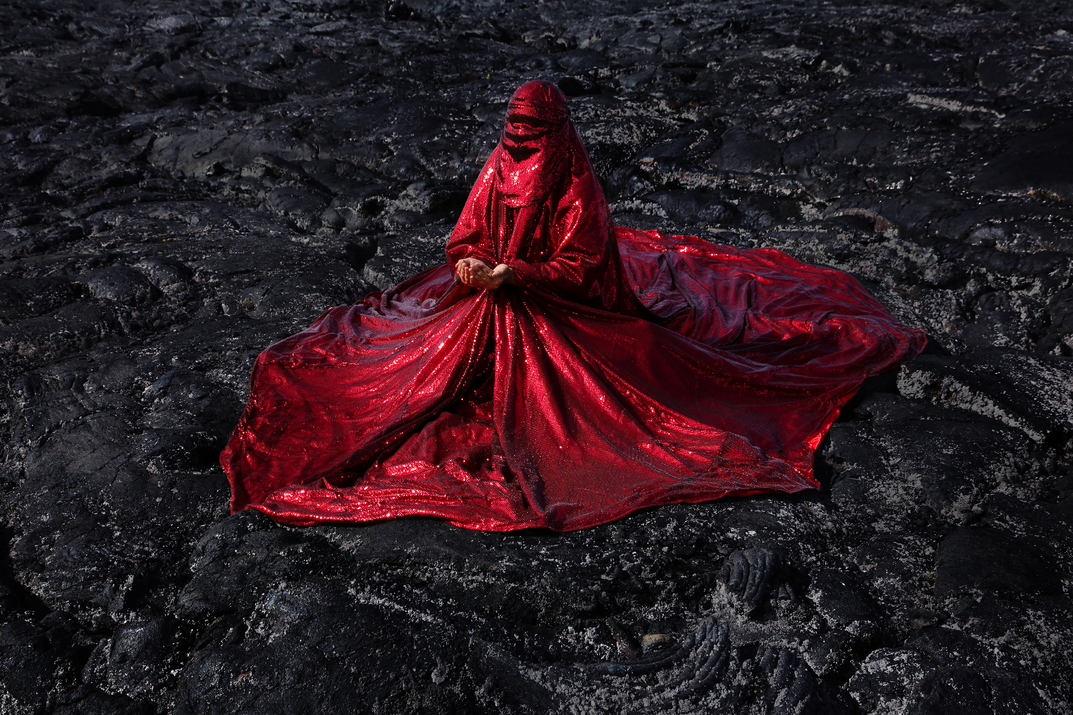 A figure draped in a flowing red glittering chador, seated gracefully on a dark, rugged lava field, creating a striking visual contrast between the vibrant fabric and the stark volcanic terrain.