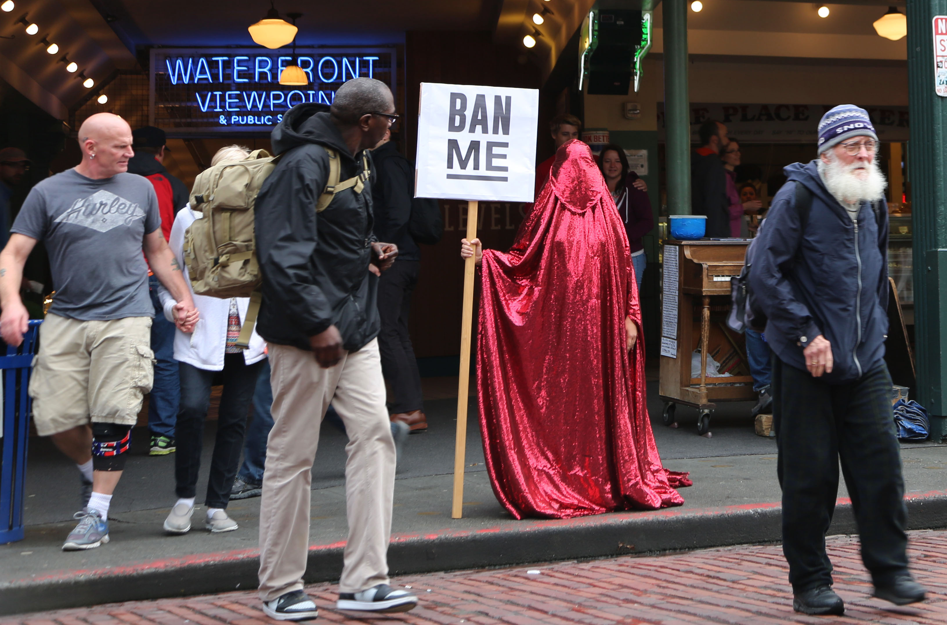 A figure draped in a red glittering chador standing on a busy street, holding a sign that reads "BAN ME," surrounded by pedestrians near a market.
