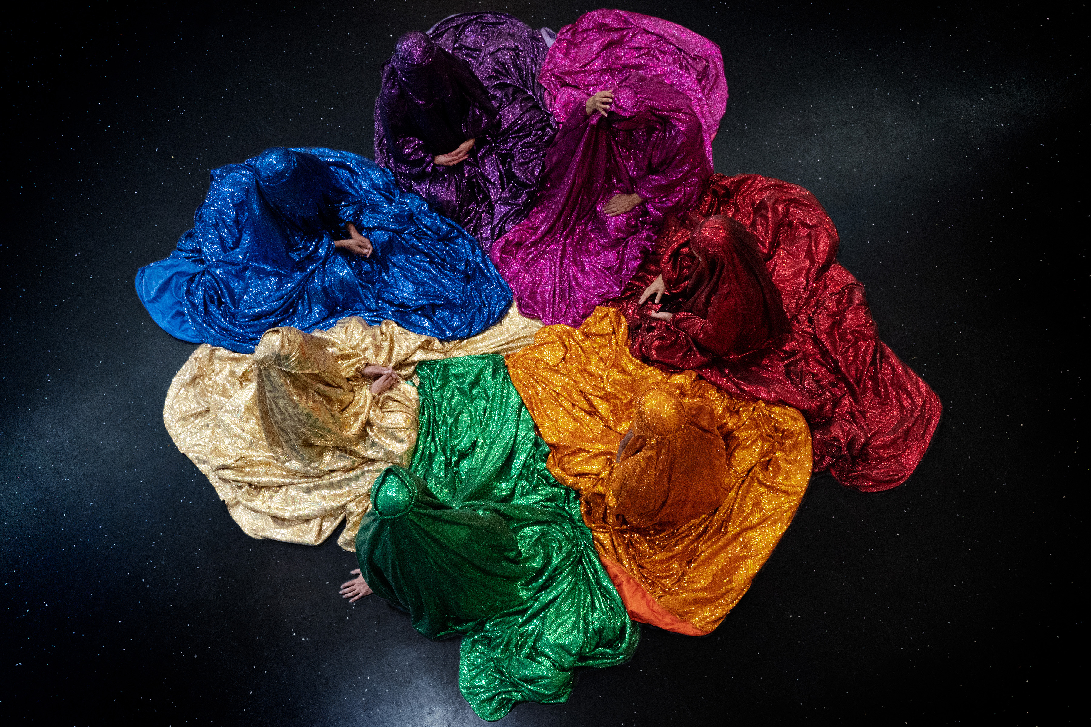 a top-down view of seven figures draped in glittering chadors of various colors—blue, purple, pink, red, orange, green, and gold—arranged in a circular formation against a dark, starry background.