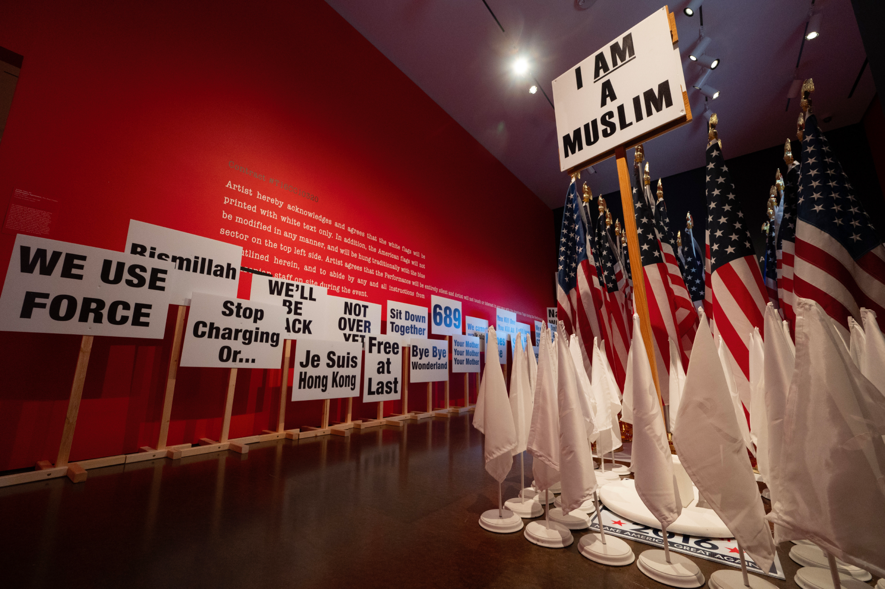 An exhibition featuring protest-style signs with messages like "I AM A MUSLIM," "WE USE FORCE," and "Je Suis Hong Kong," surrounded by rows of white flags and American flags, set against a red wall with text.