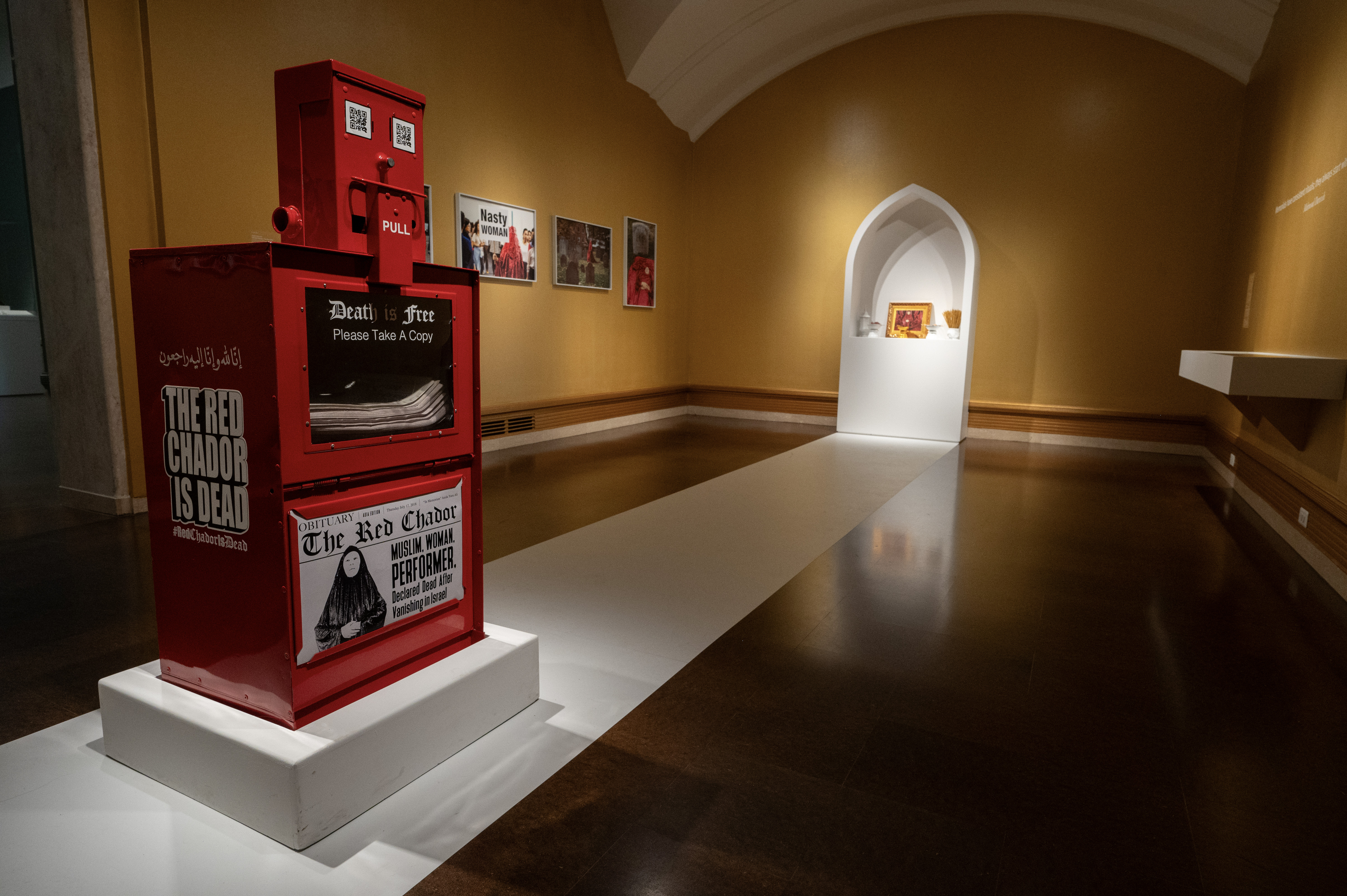 A red newspaper dispenser labeled "The Red Chador Is Dead" with an obituary display, situated in a gallery space with framed artwork on the walls and a small illuminated alcove in the background.