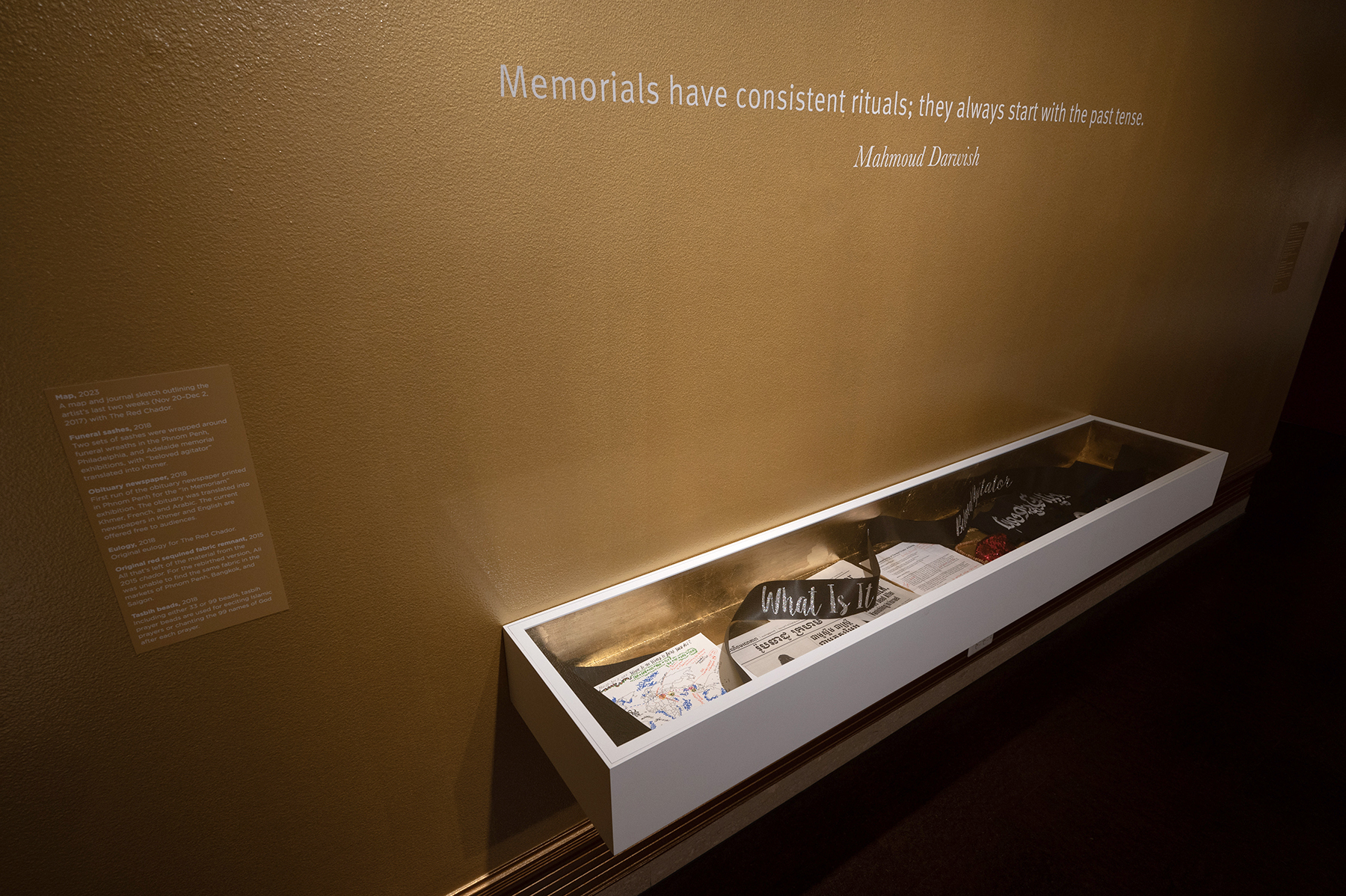  a wall-mounted display case containing various memorial-related artifacts, with a quote by Mahmoud Darwish above stating, "Memorials have consistent rituals; they always start with the past tense."