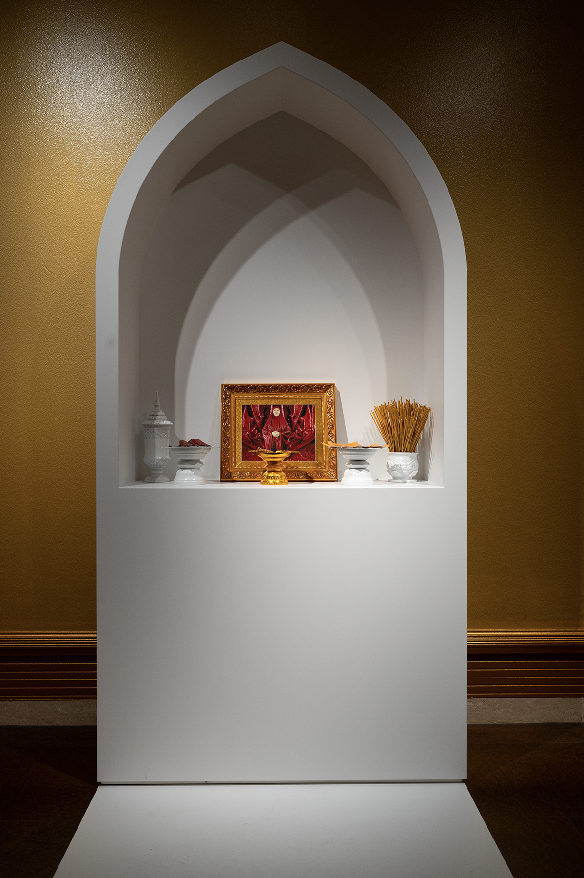 A white, arched altar-like structure displaying a framed artwork, incense sticks, and ceremonial objects arranged in a symmetrical manner.