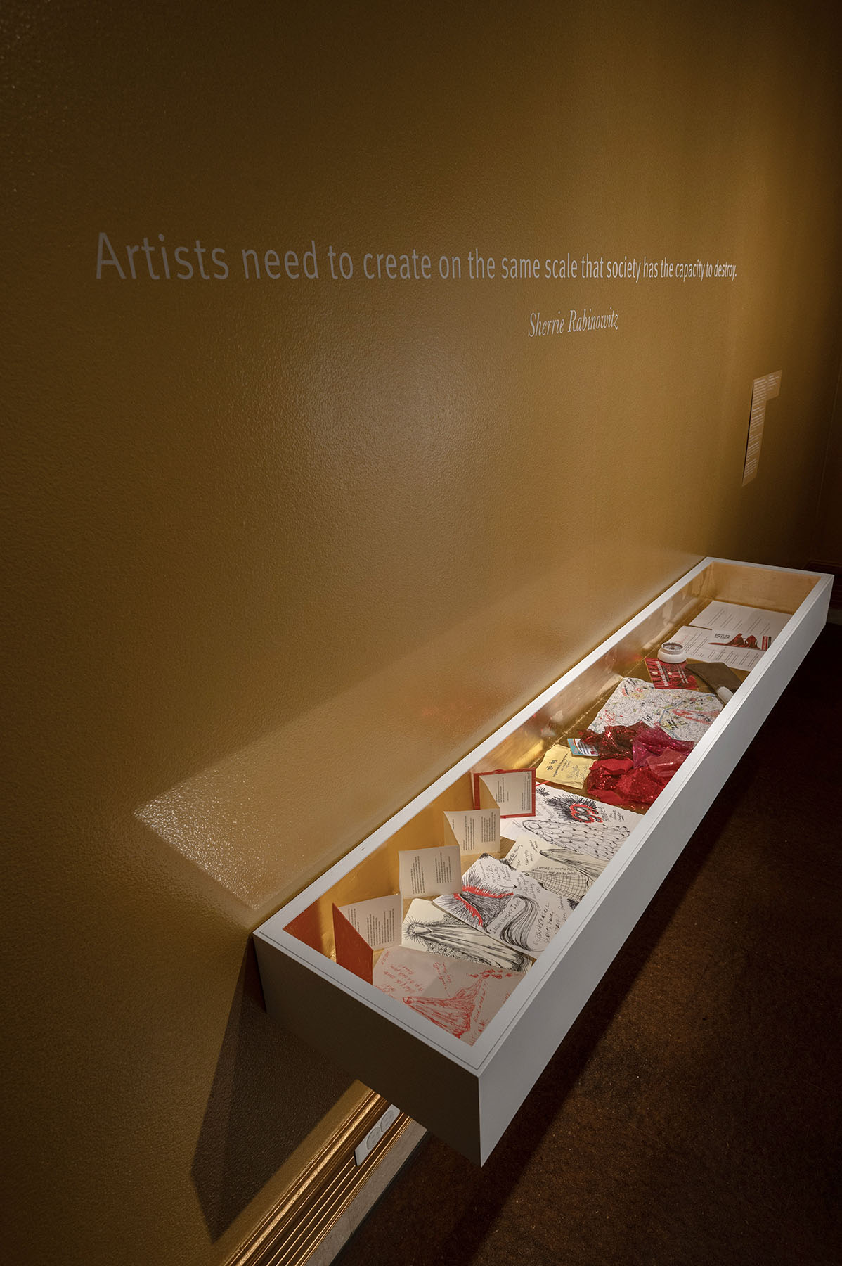 A wall-mounted display case containing artistic artifacts and handwritten notes, with a quote by Sherrie Rabinowitz above stating, "Artists need to create on the same scale that society has the capacity to destroy."