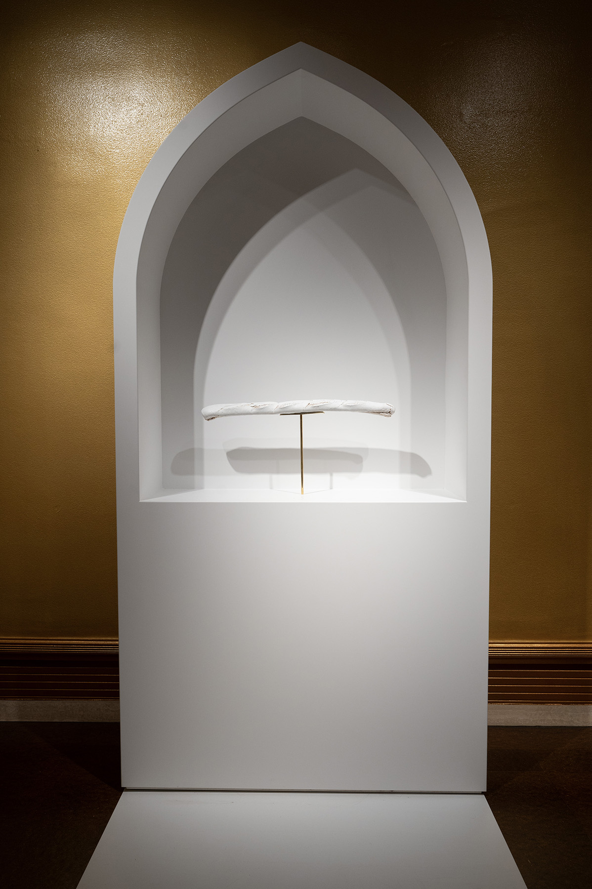 A white, arched altar-like structure displaying a single white baguette, mounted on a thin gold stand.