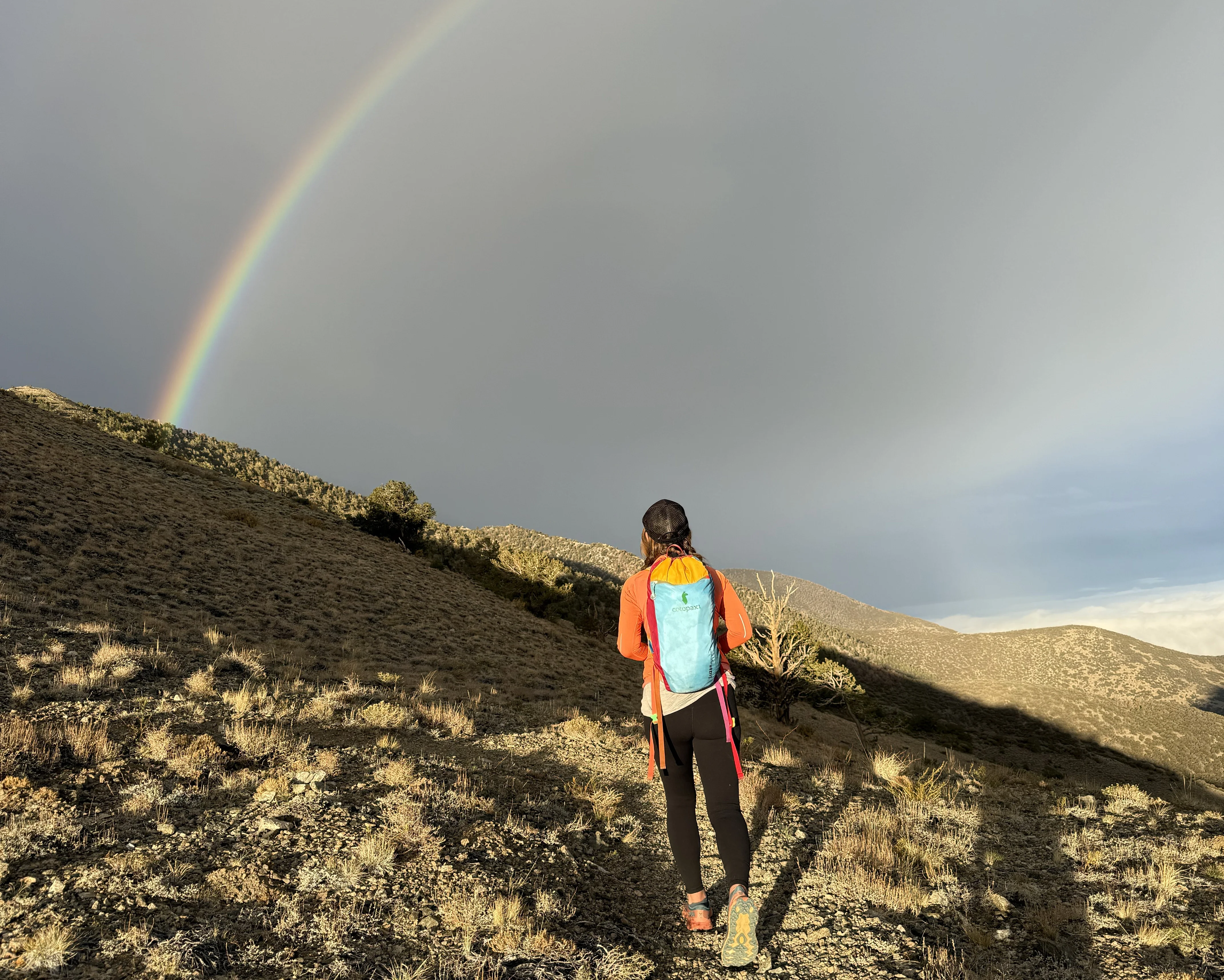 A woman in an orange hoodie with a blue backpack gazes upon a rainbow forming in the horizon.