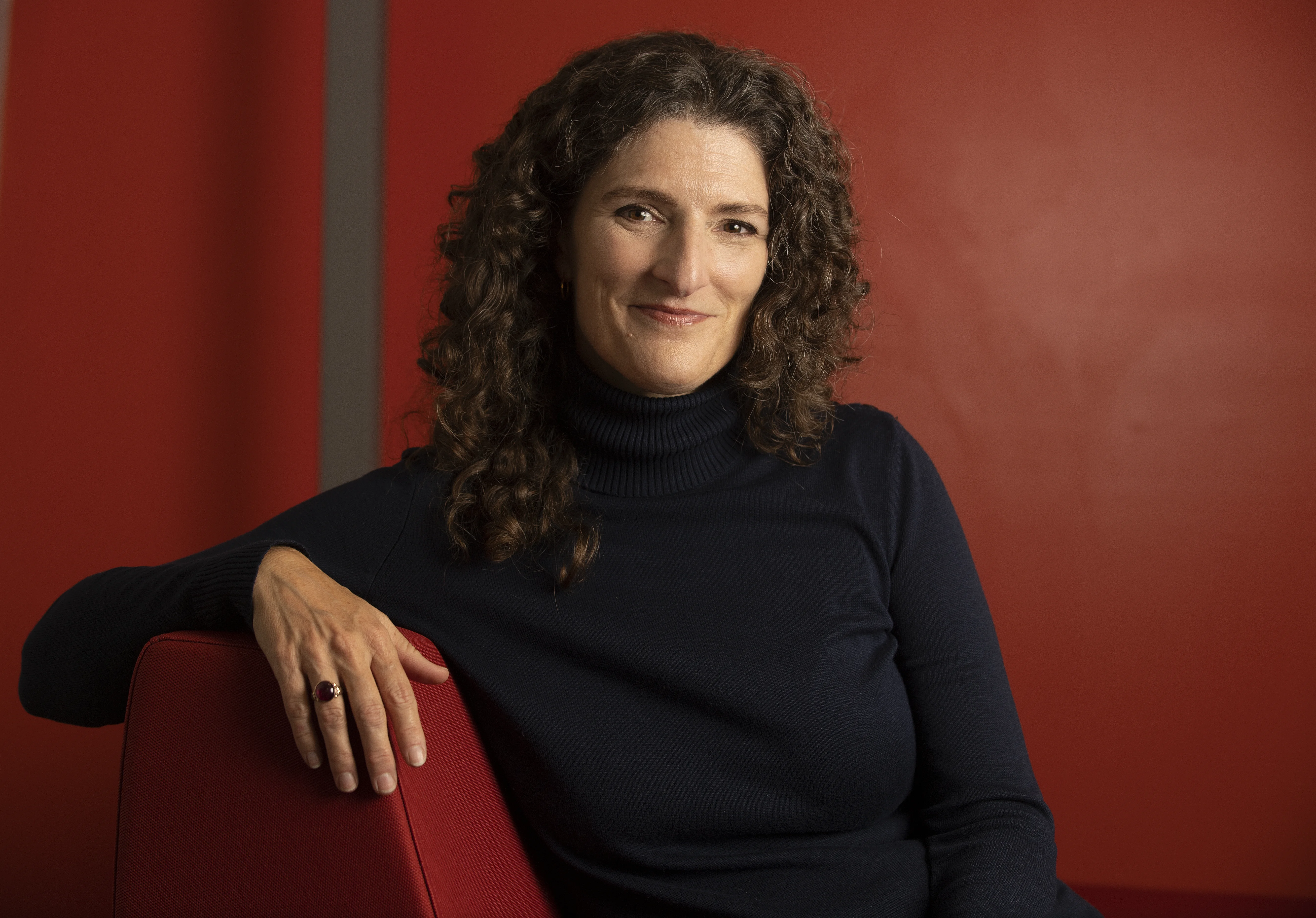 A woman with brunette curly hair rests on a chair posing for the camera with a dark red background.