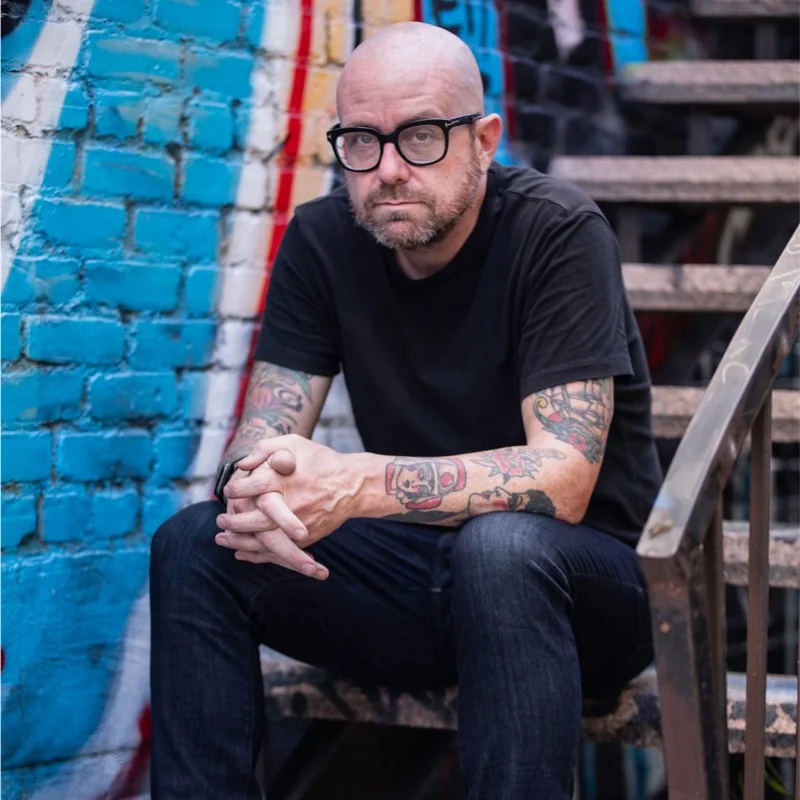 A bald man with glasses and multiple arm tattoos sits on a staircase with his arms resting over his knees.