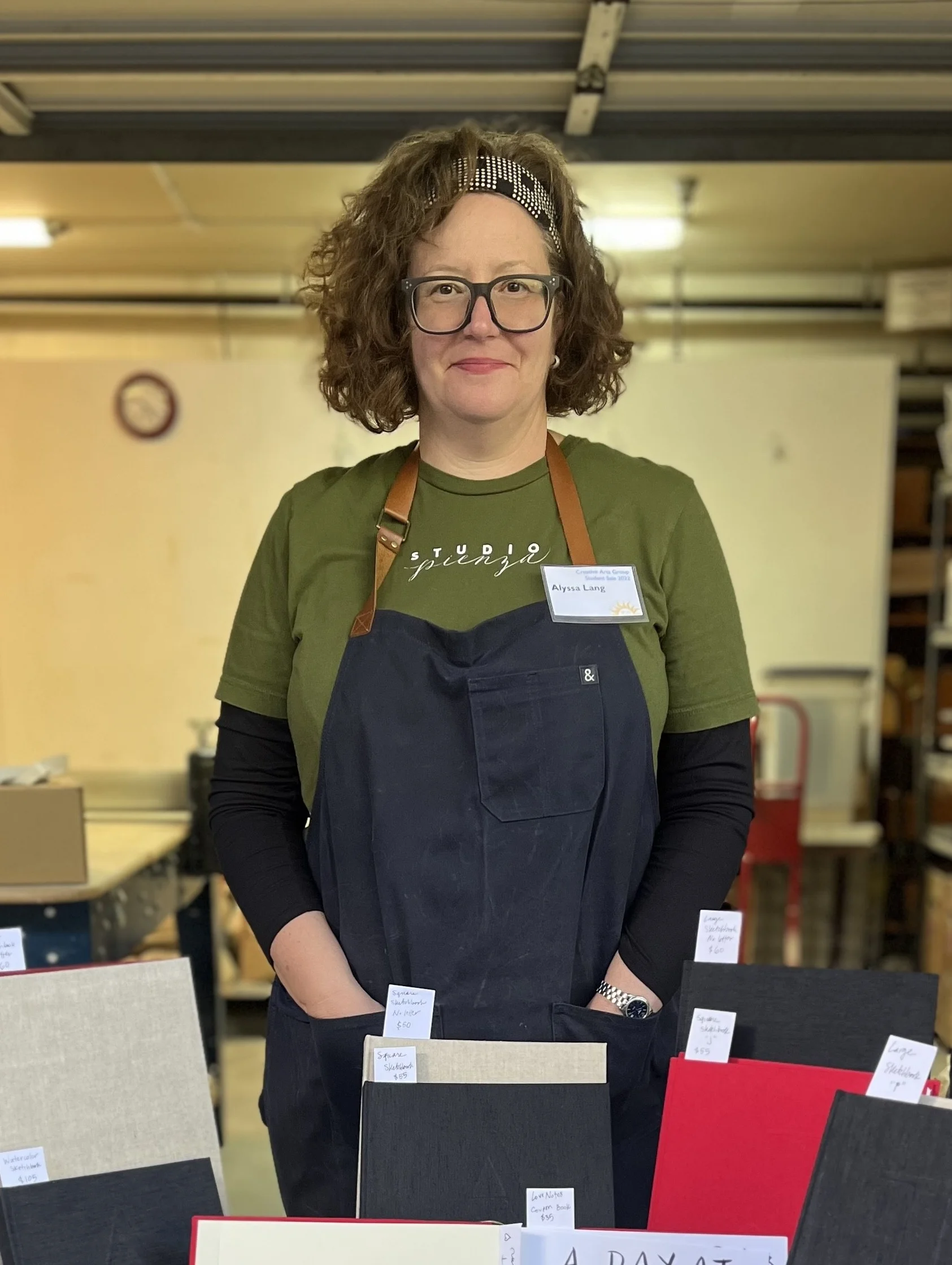 A woman with glasses wearing a navy smock smiles at the camera while in an art studio.