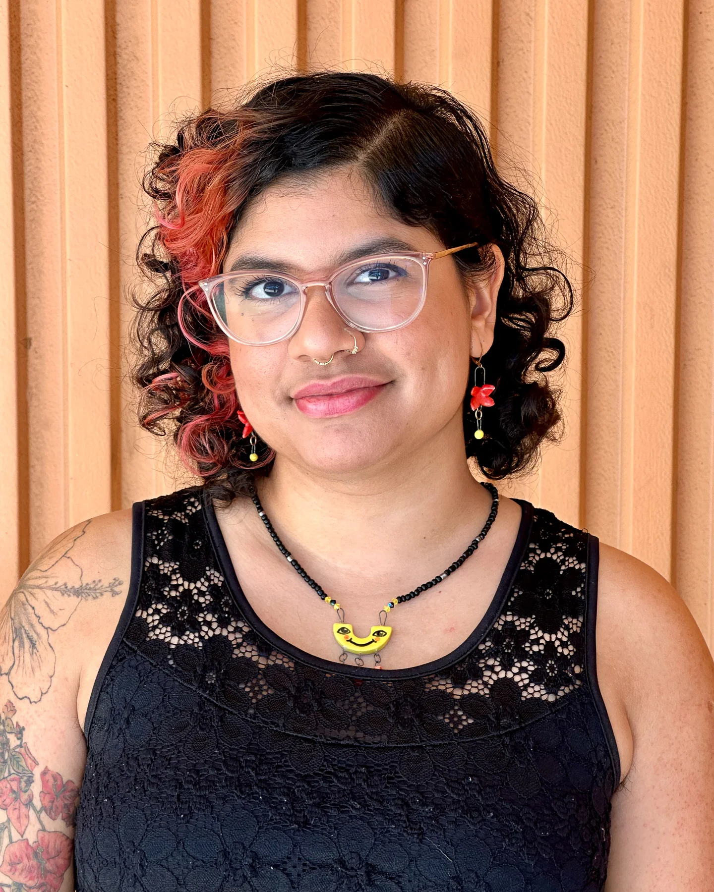 A woman with glasses, nose piercing and a red dye streak in her hair smiles at the camera.