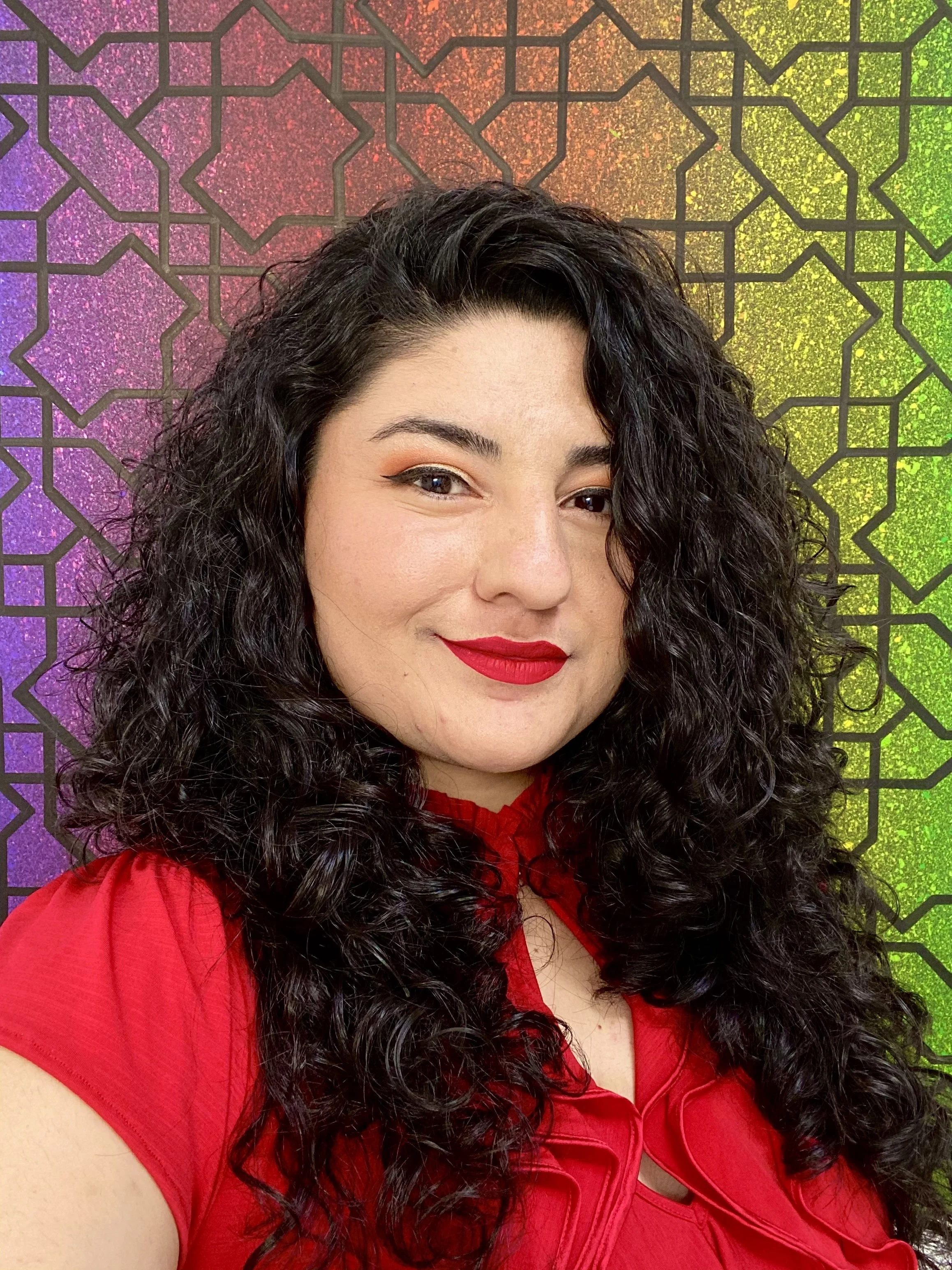 Woman in a red shirt with dark long curly hair smiles at the camera with a chroma frosted glass background.