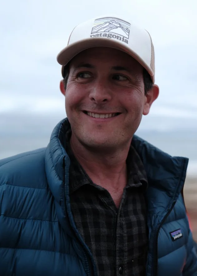 A man stands in the outdoor wearing a hat, puffer jacket and flannel.