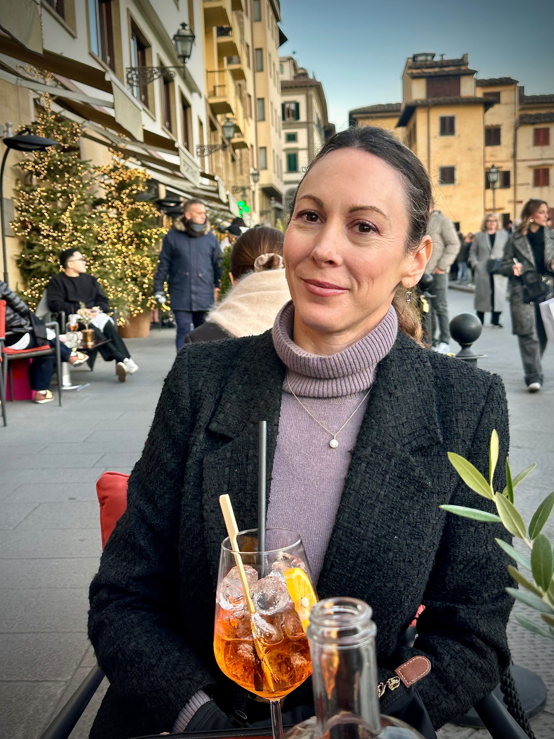Woman with hair tied back sitting in a public square with a drink in front of her.
