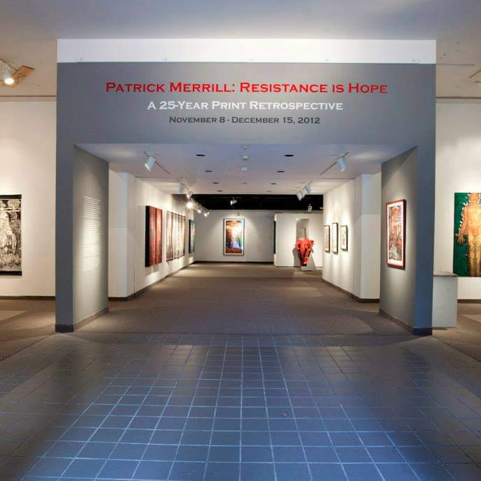 Entrance of gallery with title wall painted grey. Red, white, and dark grey text on title wall reads: Patrick Merill: Resistance is Hope A 25-year print retrospective. November 8- December 15, 2012