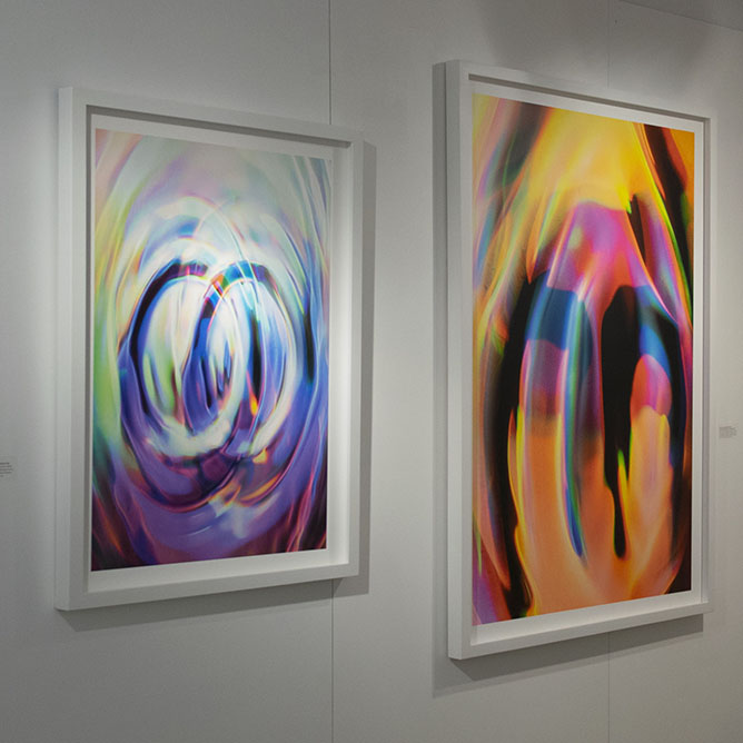 Image of two rectangular artworks mounted on gallery wall. the artwork on the left is a bit smaller. The both have images of abstract colorful ripple-like patterns.