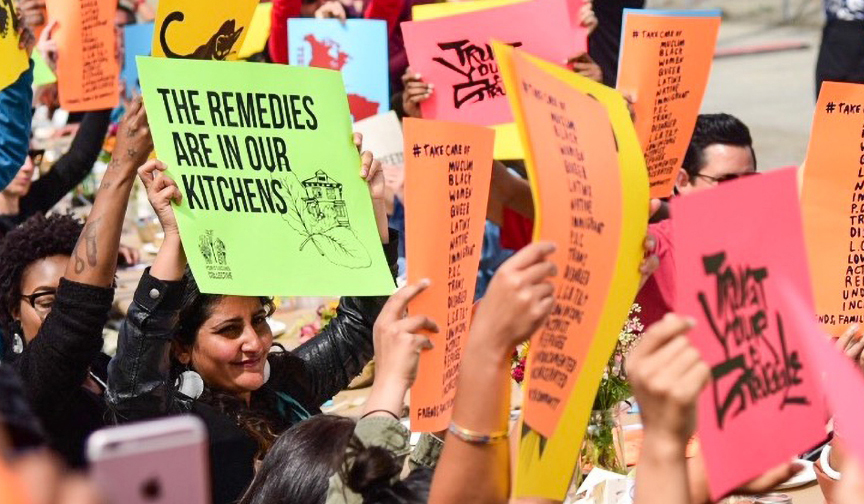  Image of group of people holding colorful signs. One sign reads “the remedies are in our kitchens” 
