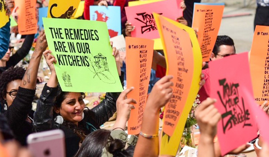 Image of group of people holding colorful signs. One sign reads “the remedies are in our kitchens”