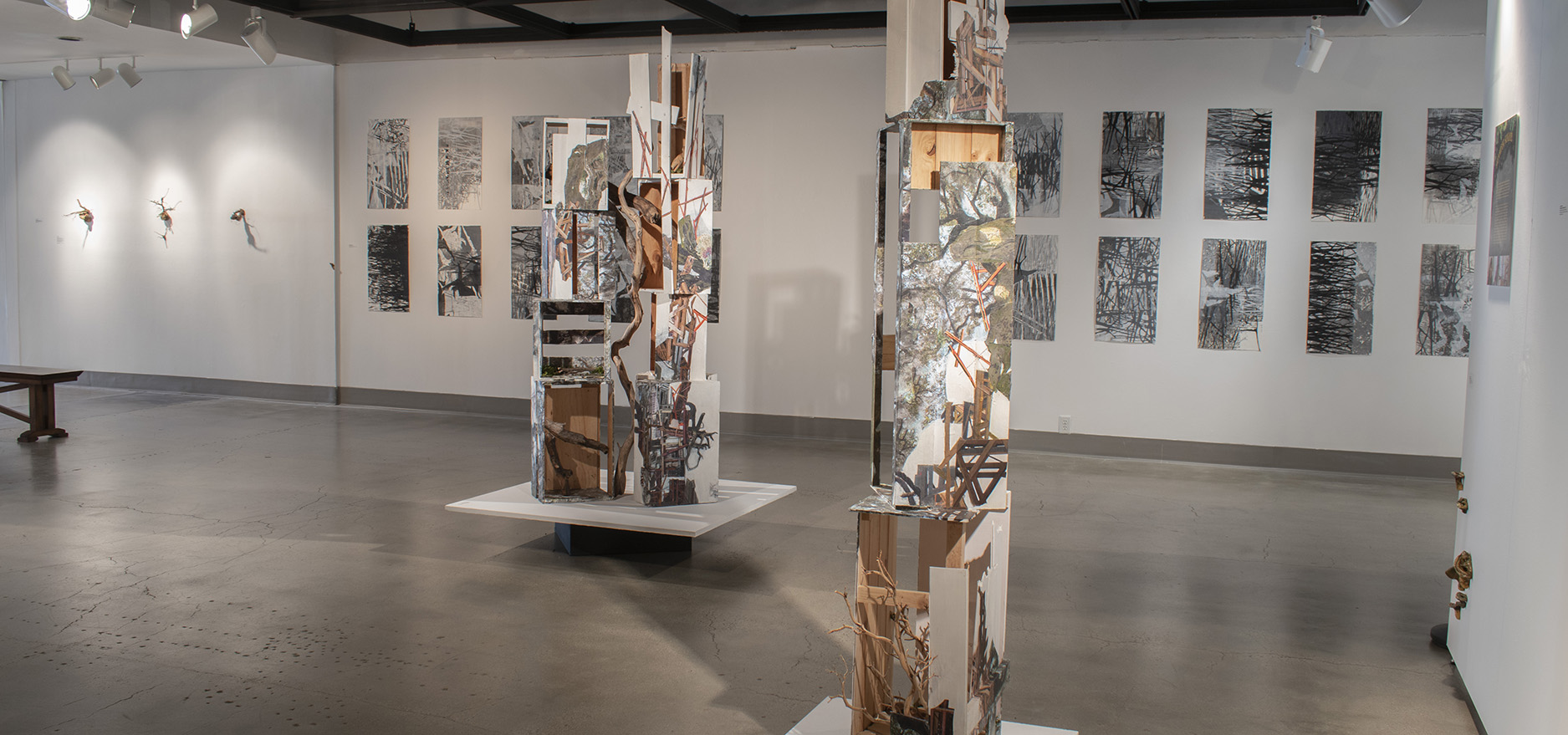 image of gallery space, sculptures of wooden crates with collages on them. Black and white rectangle artworks mounted on walls.
