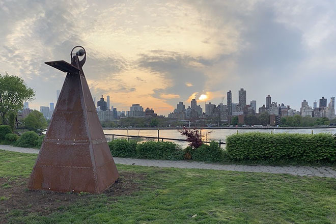 Metal, pyramid sculpture in grassy area near river that is across from a city