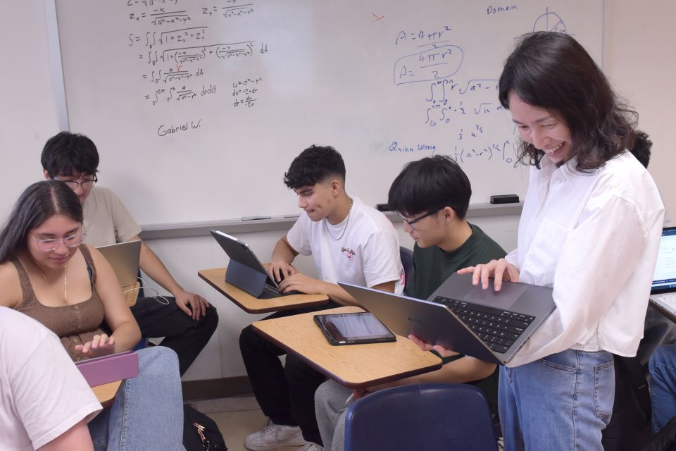 A faculty member works with students in the classroom. All are holding computers and working on something