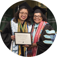 Student at a graduation posing, holding a diploma and standing next to faculty