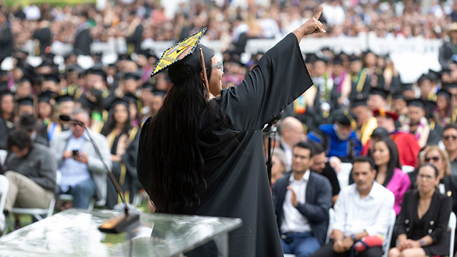 Female graduate raises hand to celebrate on the commencement stage while facing the crowd.