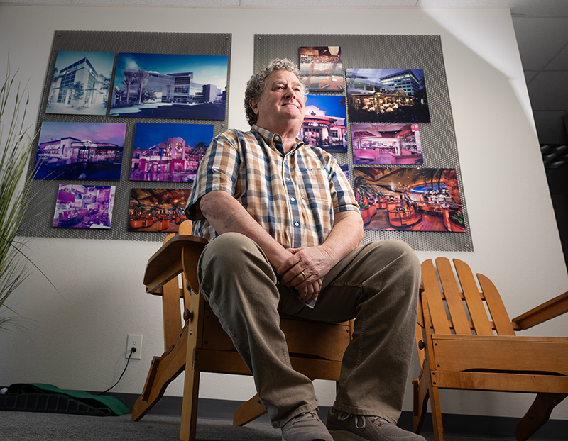 Mike Riley sits on a chair in front of a wall that holds several of his designs.