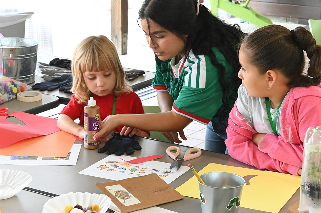 A female student instructor works with two girls at a table on a craft. 
