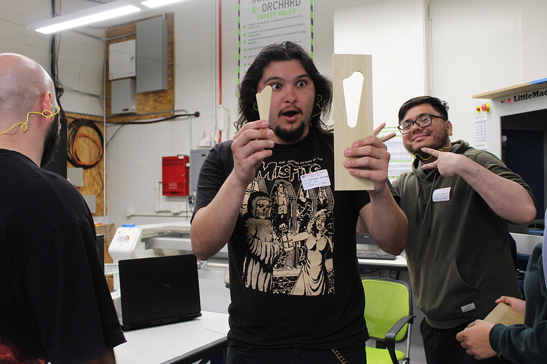 male student looks surprised while holding up a piece of wood he carved for the daxophone.