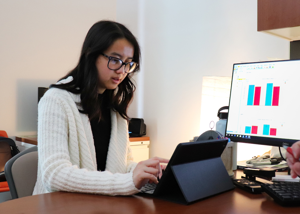 Image of Kelly Tran in front of a monitor displaying bar graphs, while she's looking down and using her tablet