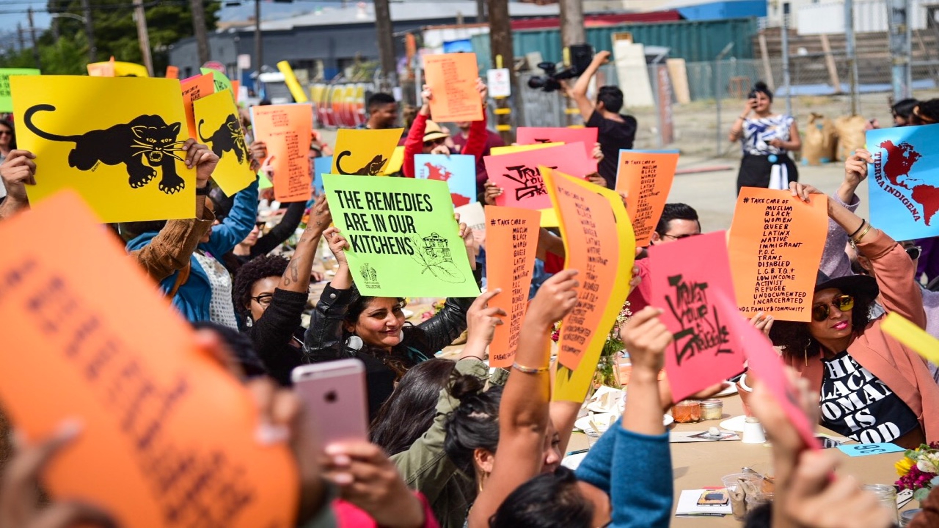 People's Kitchen Collective Free Breakfast Program at Life is Living event with large group of participants gathered in outdoor assembly holding and waiving signs and multicolor event flyers.