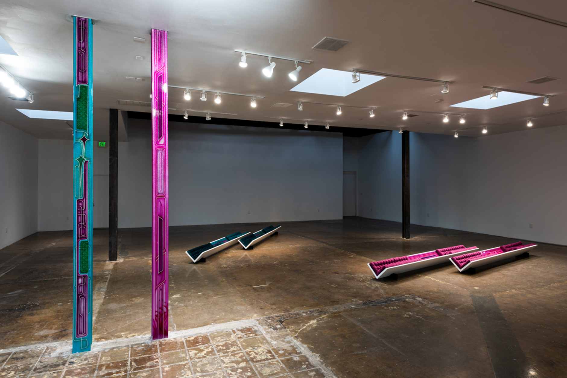 The photo is of Some Reach While Others Clap, 2020 of a bare gallery room with white walls and concrete floors. In dim lighting there are two pillars in the foreground, the left one is bright turquoise with pink designs, immediately to the right is a thin pillar that is bright pink with turquoise designs. 2 sets of 2 angle beams, one set in turquoise and the other in pink, are laying on the ground in the background stretching across the floor. All are painted in glitter paint and upholstered in matching velvet fabric referencing low-rider cars.