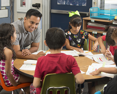 A male teacher sitting at a table surrounded by young students