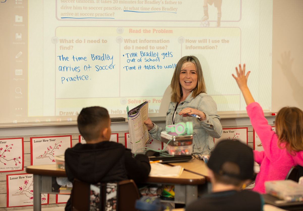 A teacher stands in front of the whiteboard while young kids raise their hands