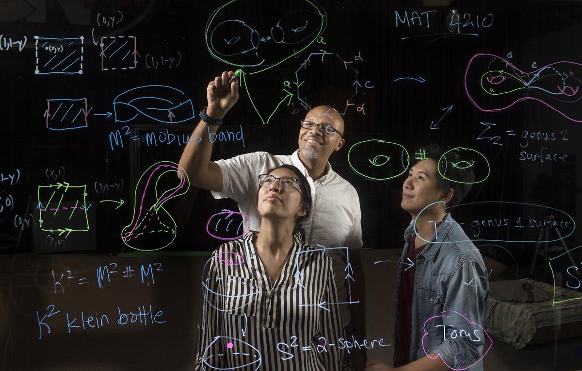 Professor and students solve equations on a transparent board. 