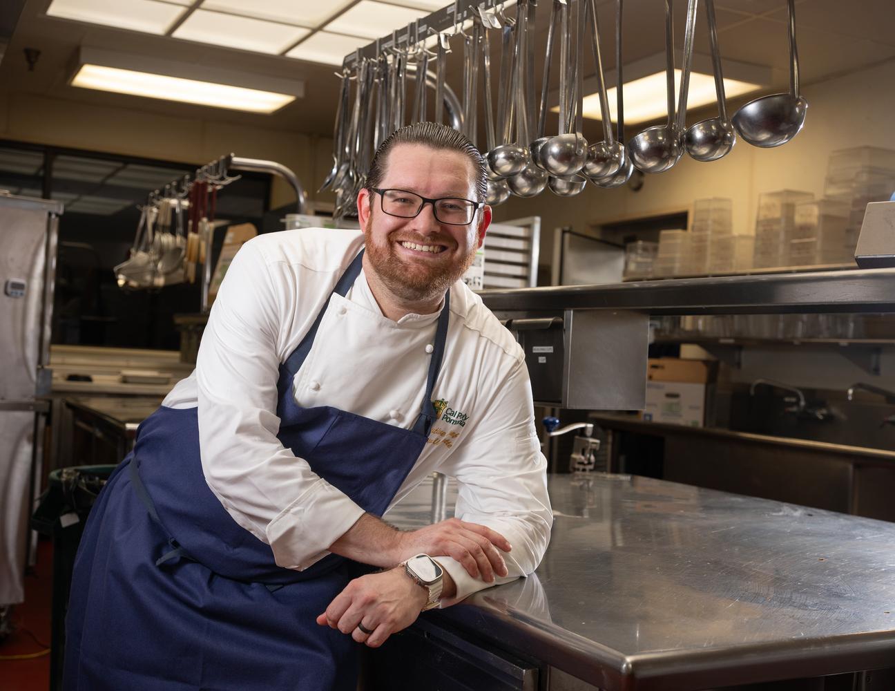 Chef Matt leaning on a silver kitchen table in his apron and white uniform.