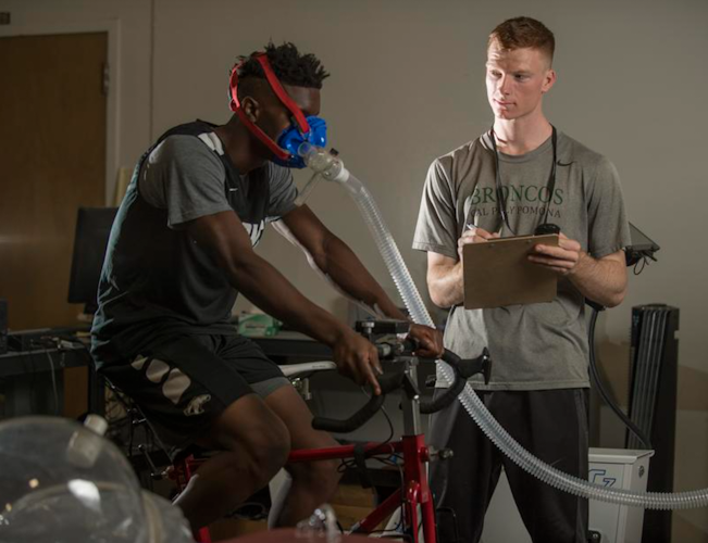 One student using an exercise bike wearing a mask to measure oxygen output while cycling, a second student stands beside the exercise bike taking notes.