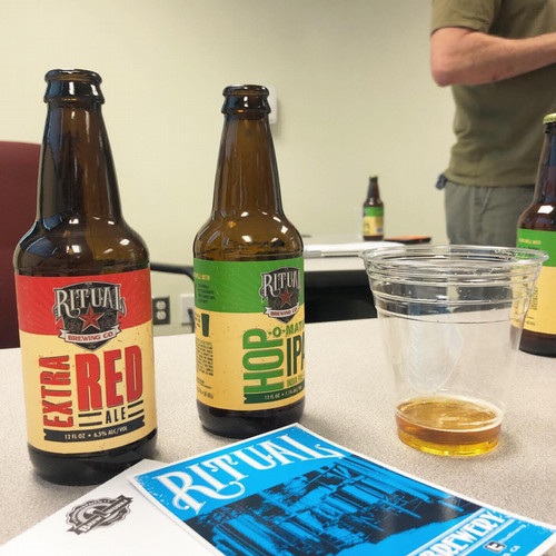 Two bottles of a beverage behind a flyer, next to a plastic cup with a small serving of the beverage