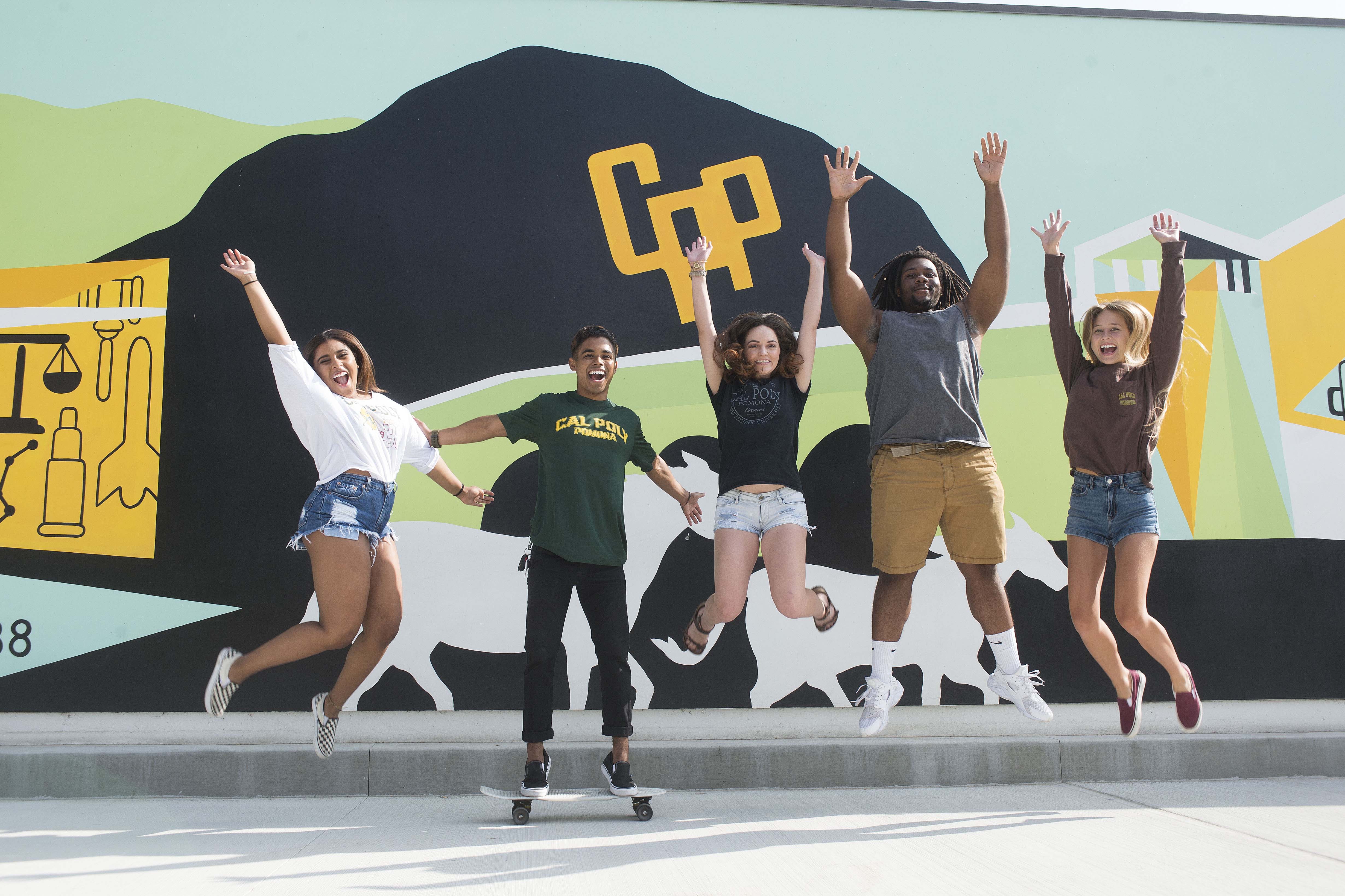 A group of 5 students jumping in front of the mural by the new parking structure