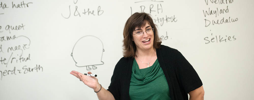 A white woman teaches a course. There is writing on the whiteboard behind her