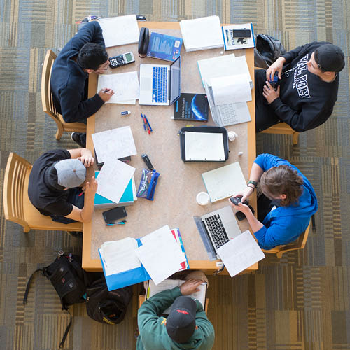 An overhead view of a group of male students studying. Laptops, phones and homework are spread across the table