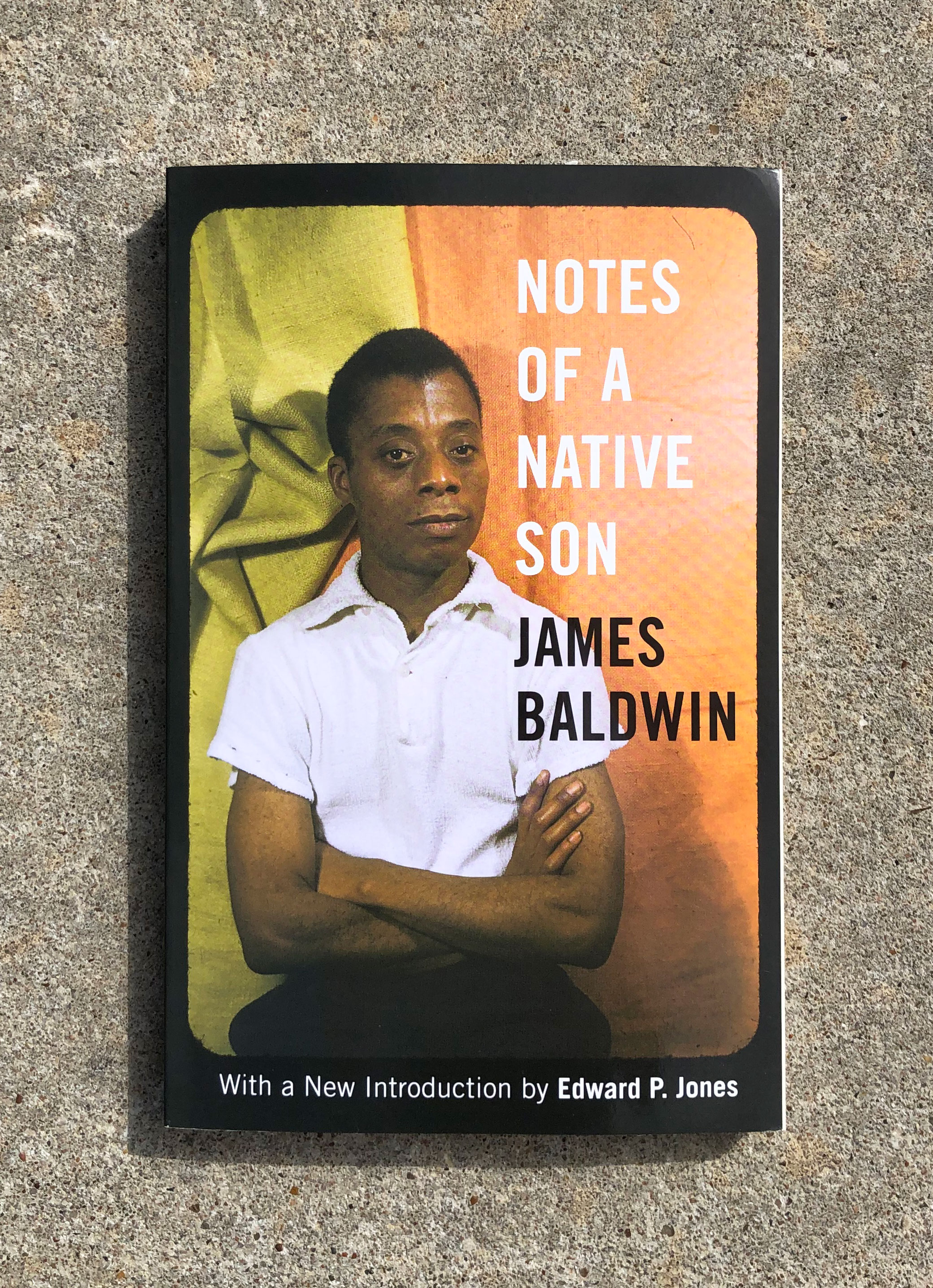 The cover of a book against a concrete background with a photo of author James Baldwin sitting in front of an orange and green background. Next to his image in white text it reads, "Notes of a Native Son" and then in black text "James Baldwin". At the bottom within in a black border surrounding the image, in white text, it reads, "with a new introduction by Edward P. Jones"