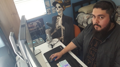 Student & podcaster Chris Vasquez sits at his computer desk wearing headphones and looking at the camera. Also in the room is his prop skeleton Bernie, wearing a baseball cap and seemingly talking into a desktop microphone.