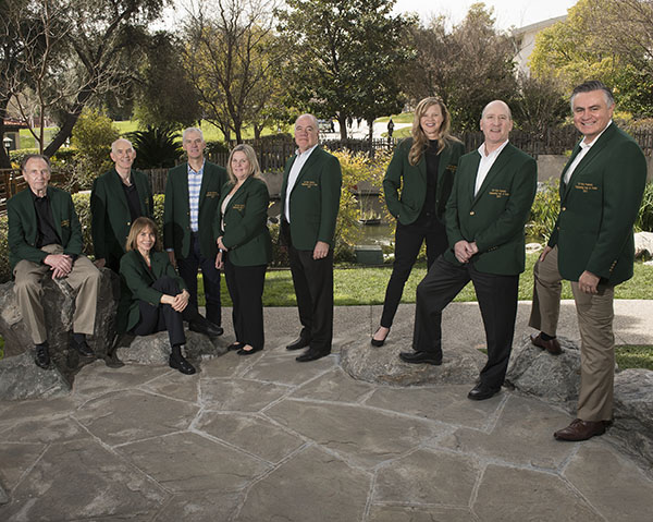 Nine individuals, seven men and two women, all with green blazers and posing as a group.