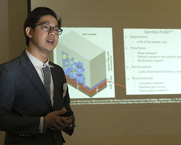 A young Asian man presenting a a technical engineering PowerPoint slide. The text in the slide is illegible.