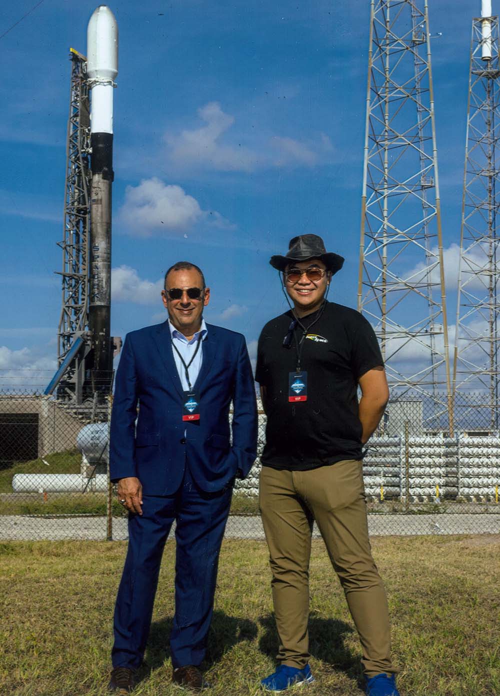 Two men standing and smiling in front of a rocket launch site.